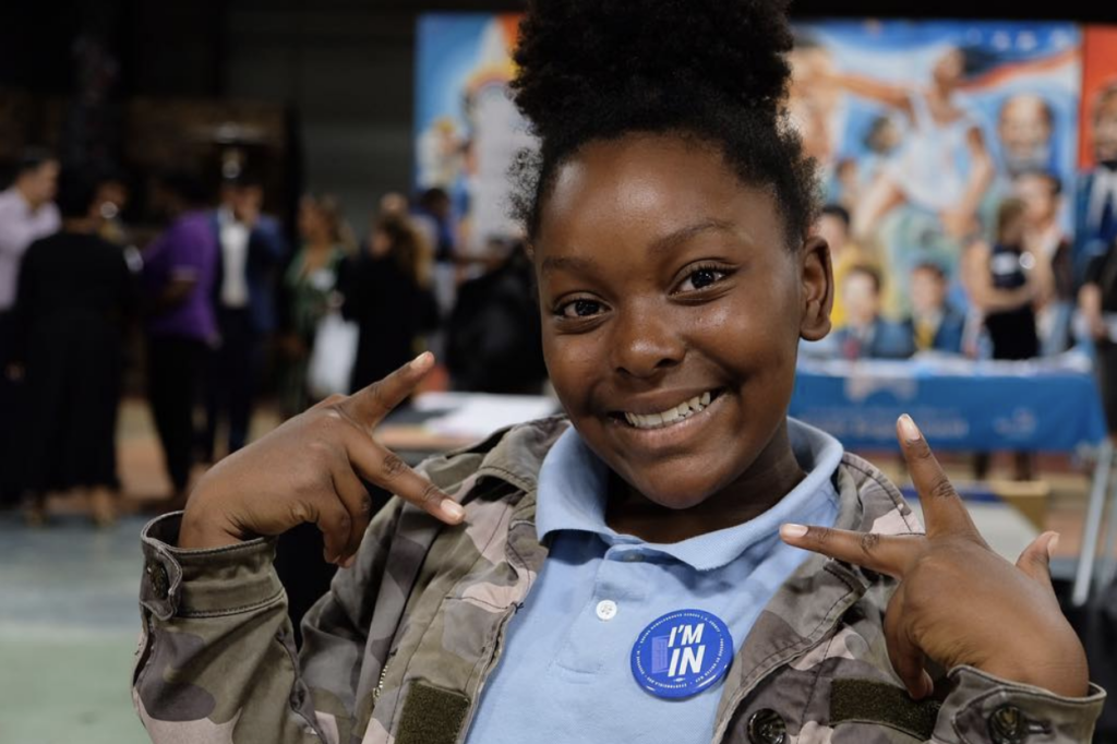 A young girl wearing an Everyone In pin smiles and holds up peace signs on both hands